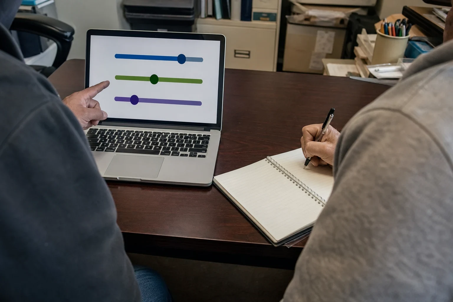 Over-the-shoulder view of two people at an office desk, one pointing to slider options on a laptop screen while the other writes notes in a spiral notebook